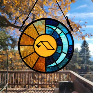 May include: A stained glass suncatcher with a yellow, orange, and blue design. The suncatcher features a stylized letter "A" in the center.