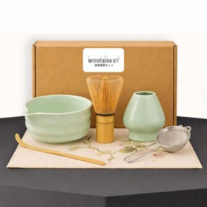 May include: A matcha whisk set includes a light green ceramic bowl, a bamboo whisk, a small vase, a bamboo scoop, and a stainless steel strainer. The set is arranged on a floral patterned mat, with a cardboard box in the background. The box has a label that says "Matcha Whisk Set".