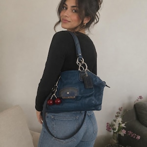 May include: A dark blue leather handbag featuring a silver-toned metal ring attachment on the strap, a rectangular flap pocket with a snap closure, and a dangling red cherry charm. The bag includes a leather luggage tag and a zipper closure.