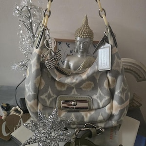 May include: A grey and cream patterned hobo handbag featuring a silver-tone turn-lock closure on the front pocket, a single shoulder strap with cream-colored ring accents, a silver metallic luggage tag, and a dangling charm with two crystal-encrusted cherries.