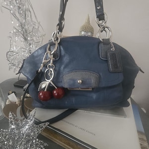 May include: A blue leather handbag featuring a front flap pocket with a textured gray accent, silver-tone hardware, and a dangling charm with two red cherries. The bag includes a dark gray leather handle and a rectangular leather Coach brand tag.