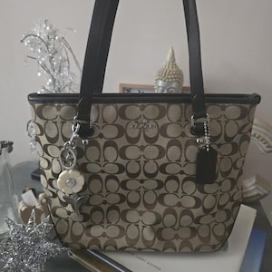May include: A beige and brown Coach tote bag featuring the signature C monogram pattern, dark brown leather shoulder straps, silver-tone hardware, a black leather hangtag, and a white flower-shaped charm attached to the front metal ring.