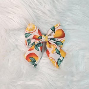 May include: A large, fabric hair bow featuring a citrus fruit pattern. The bow has a light pink background with orange slices, whole oranges, green leaves, and white flowers. The bow is centered on a fluffy white surface.