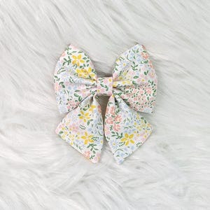 May include: A large, floral hair bow featuring a mix of pastel colors. The bow is made of fabric with a pattern of small flowers in yellow, pink, and blue on a white background. The bow is tied in the center with a matching fabric piece.