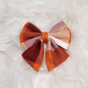 May include: A fabric hair bow featuring a plaid pattern in shades of burnt orange, deep maroon, and white. The bow is constructed with folded fabric sections and a solid orange center band, creating a multi-layered appearance with distinct diagonal lines.