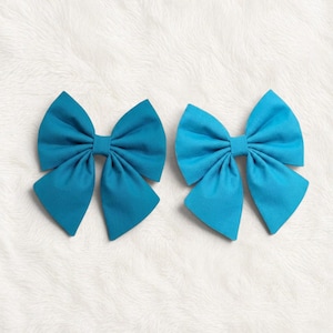 May include: Two teal fabric hair bows. Each bow is a symmetrical design with a central knot and four flared loops. The bows are set against a white, fluffy background.