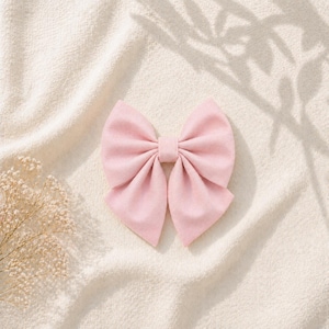 May include: A light pink fabric bow, centered on a cream-colored textured surface. The bow is neatly tied with symmetrical loops and tails. Soft shadows from foliage are cast across the background, adding a delicate touch.