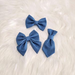 May include: Three denim blue bow ties and a necktie are arranged on a white, fluffy surface. The bow ties vary in size, with one smaller and two larger. The necktie is a solid, matching blue. These accessories are suitable for pets or people.