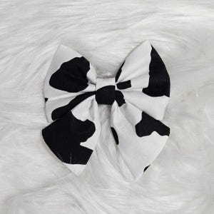 May include: A white and black cow print bow. The bow is made of fabric and has a large, rounded shape.