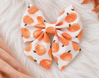 May include: A fabric hair bow featuring a repeating pattern of orange pumpkins on a white background. The bow is constructed with multiple folded loops and a central knot, showcasing a seasonal design with a soft, structured appearance.