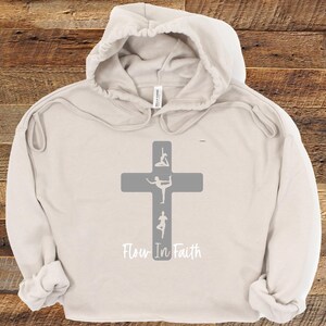 May include: A beige hooded sweatshirt with a gray cross and the text "Flow In Faith". The cross has four yoga poses on it.