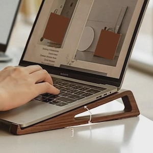 May include: A wooden laptop stand with an adjustable angle. The stand is made of dark brown wood and supports a silver laptop. The laptop screen displays a webpage with brown shopping bag graphics. Another laptop is visible in the background.