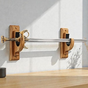 Puede incluir: Expositor decorativo de espada con una hoja plateada y un mango dorado ornamentado. La espada está montada en dos soportes de pared de madera con un acabado natural. Los soportes están diseñados para sujetar la espada de forma segura.