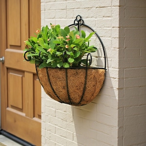 May include: A black metal hanging basket filled with green plants and small pink flowers. The basket has a brown, woven interior and is mounted on a white brick wall near a wooden door.