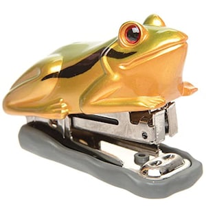 May include: A gold frog-shaped stapler with red eyes. The stapler has a gray base and a silver metal staple mechanism.