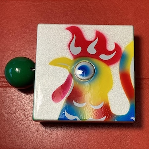May include: A square, decorative box with a colorful rooster design. The rooster features red, yellow, blue, and white accents. A green, spherical knob is attached to the side of the box.