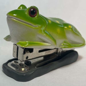 May include: A green frog-shaped stapler with red eyes and a silver stapling mechanism. The frog's body is a glossy green with white and yellow accents. The base of the stapler is black.