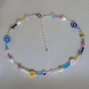 May include: A colorful beaded necklace with small flower charms in blue, yellow, and pink. The necklace is made with white, blue, yellow, and pink beads.