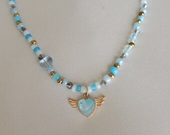 Puede incluir: Un delicado collar de cuentas azules y doradas con un dije de corazón alado. El corazón es azul claro con un borde dorado y alas.