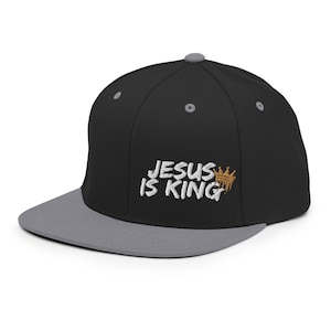 Könnte beinhalten: Schwarze und graue Baseballkappe mit einem weißen gestickten Design, das "JESUS IS KING" mit einer goldenen Krone über dem Wort "KING" sagt.