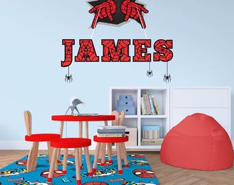 Puede incluir: Una alfombra roja y azul con temática de Spider-Man con una mesa y dos sillas. La mesa y las sillas son de madera y tienen un acabado rojo. La alfombra tiene un fondo azul con gráficos de Spider-Man rojos y blancos. La pared detrás de la mesa tiene una calcomanía de pared con temática de Spider-Man con el nombre "JAMES" escrito en letras rojas.