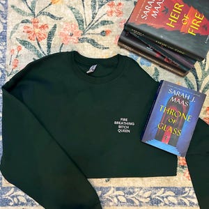 May include: Dark green sweatshirt with the text "FIRE BREATHING BITCH QUEEN" in white. Several books by Sarah J. Maas are stacked nearby, including "Heir of Fire" and "Throne of Glass". The items are on a floral patterned rug.