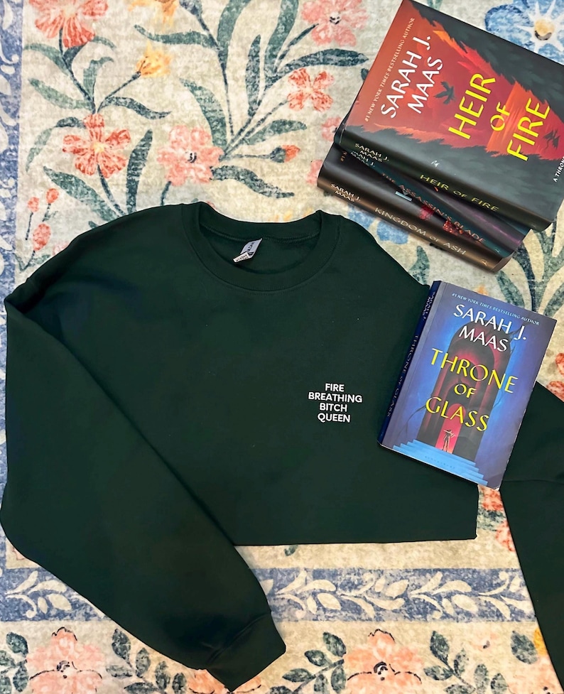 May include: Dark green sweatshirt with the text "FIRE BREATHING BITCH QUEEN" in white. Several books by Sarah J. Maas are stacked nearby, including "Heir of Fire" and "Throne of Glass". The items are on a floral patterned rug.