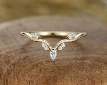 Puede incluir: Un anillo dorado con un diseño curvo. El anillo presenta un diamante en forma de pera en el centro, rodeado de diamantes redondos más pequeños. El anillo tiene una estética delicada inspirada en la naturaleza.