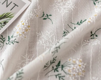 Op de afbeelding: Een doorschijnende witte stof met verticale gestructureerde strepen en een delicaat bloemenpatroon met groene stengels en kleine witte en gele bloesems. Een hoek van een witte kaart met de gedeeltelijke zwarte tekst ENT is linksboven zichtbaar.