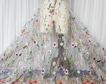 May include: A sheer, translucent fabric overlay featuring intricate, multi-colored floral embroidery. The design showcases various wildflowers, stems, and leaves in shades of red, pink, blue, yellow, and green, draped over a dress form mannequin.