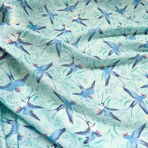 May include: Light blue fabric with a repeating pattern of blue swallows with red accents, set against a backdrop of green foliage and small white flowers. The fabric has a slightly wrinkled texture.