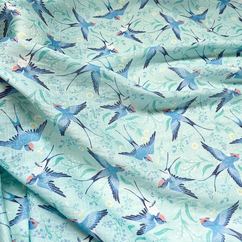 May include: Light blue fabric with a repeating pattern of blue swallows with red accents, set against a backdrop of green foliage and small white flowers. The fabric has a slightly wrinkled texture.