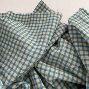 May include: Close-up of a blue and beige gingham fabric. The fabric is folded and draped, showing the pattern of intersecting lines forming a checkered design. The fabric has a soft, textured appearance, suitable for various sewing projects.