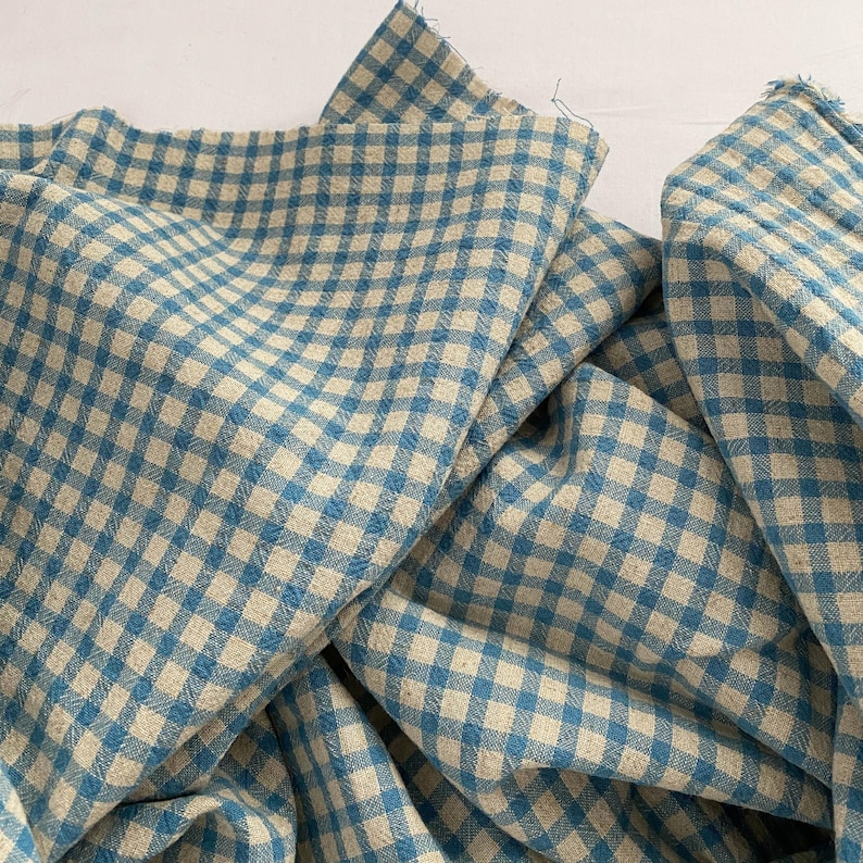 May include: Close-up of a blue and beige gingham fabric. The fabric is folded and draped, showing the pattern of intersecting lines forming a checkered design. The fabric has a soft, textured appearance, suitable for various sewing projects.