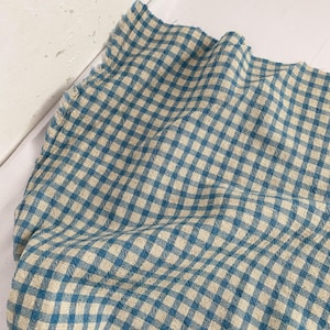May include: A close-up of a blue and beige checkered fabric. The fabric is folded and draped, showing the texture and pattern of the woven material. The pattern is a classic gingham design, with a soft, muted color palette.