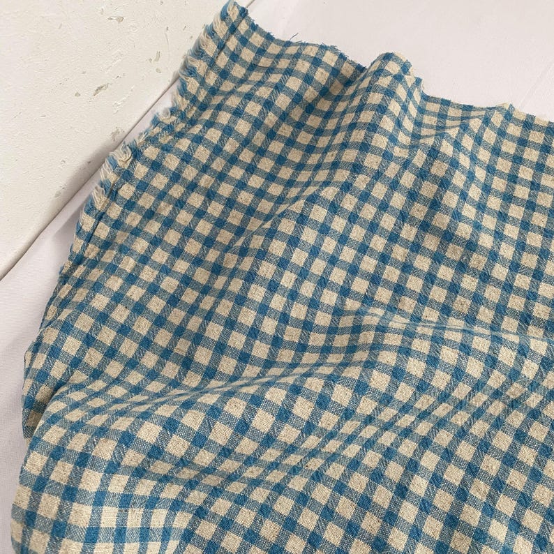 May include: A close-up of a blue and beige checkered fabric. The fabric is folded and draped, showing the texture and pattern of the woven material. The pattern is a classic gingham design, with a soft, muted color palette.