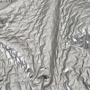 May include: Close-up of a silver quilted fabric with a diamond pattern. The fabric has a shiny, metallic appearance and is slightly wrinkled, creating shadows and highlights. The stitching is visible, adding texture.