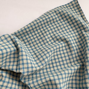 May include: A blue and white checkered fabric, likely linen or cotton, is draped across a white surface. The fabric has a classic gingham pattern, with the blue squares contrasting against the off-white background. The cloth is slightly wrinkled.