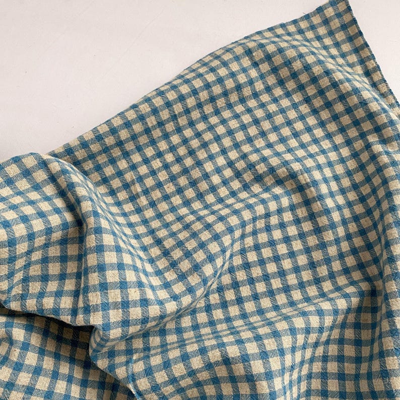 May include: A blue and white checkered fabric, likely linen or cotton, is draped across a white surface. The fabric has a classic gingham pattern, with the blue squares contrasting against the off-white background. The cloth is slightly wrinkled.