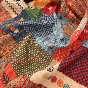 May include: A colorful patchwork fabric with various patterns and designs. The fabric features squares of different colors, including red, blue, green, and beige, with floral, geometric, and abstract motifs. The fabric is folded, creating a textured appearance.