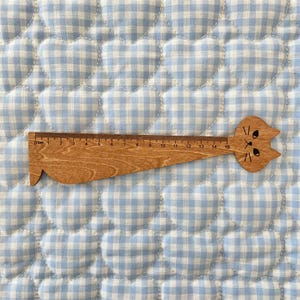 May include: A wooden ruler shaped like a cat with a long body and a feline head at the right end. The ruler features black printed markings and numbers from 0 to 15 centimetres along the top edge, with a cat face design including eyes and whiskers.