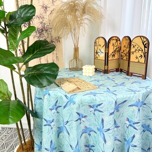 May include: A round tablecloth with a light blue background and a pattern of blue birds and flowers. A small, decorative folding screen with a bamboo design sits on the table. A plant and other decorative items are in the background.