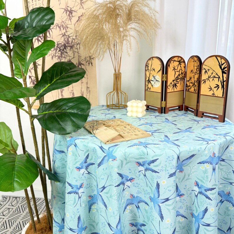 May include: A round tablecloth with a light blue background and a pattern of blue birds and flowers. A small, decorative folding screen with a bamboo design sits on the table. A plant and other decorative items are in the background.