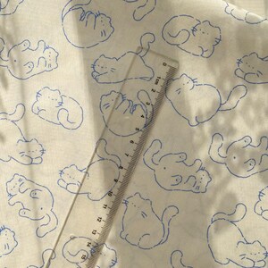 May include: White fabric with a repeating pattern of blue cartoon cats in various poses. A clear ruler with centimeter markings is placed diagonally across the fabric, measuring up to 15 cm.