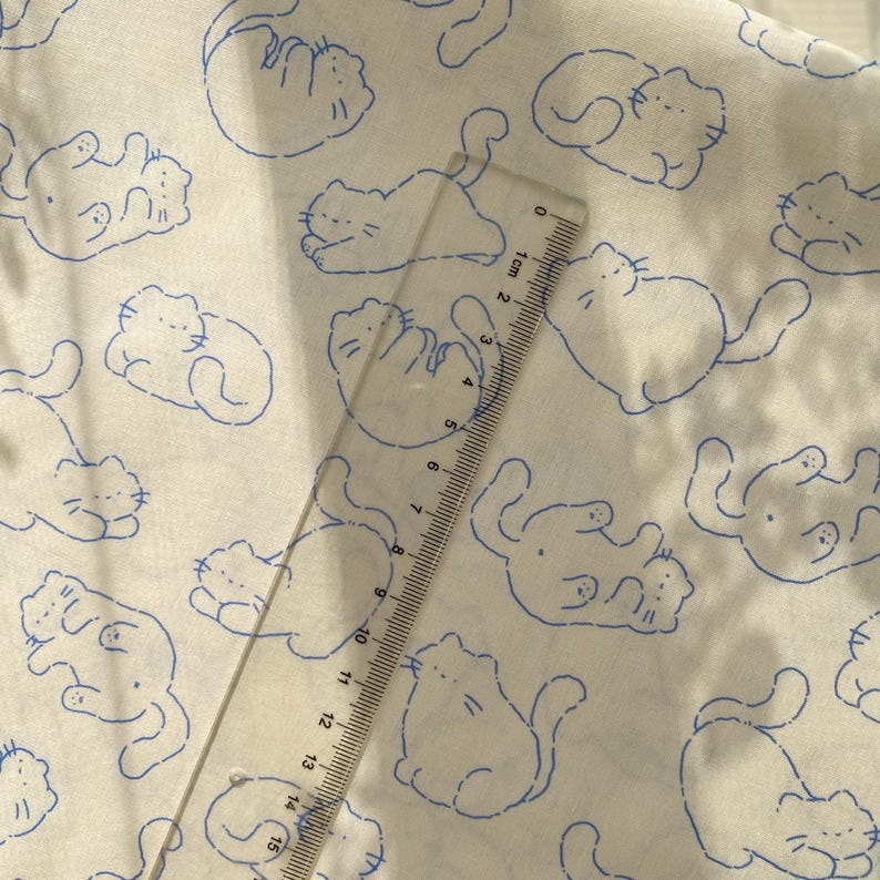 May include: White fabric with a repeating pattern of blue cartoon cats in various poses. A clear ruler with centimeter markings is placed diagonally across the fabric, measuring up to 15 cm.