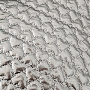 May include: Close-up of a silver quilted fabric with a repeating pattern of heart-shaped designs. The stitching is visible, creating a textured surface. The fabric has a slight sheen, reflecting light and adding depth to the design.