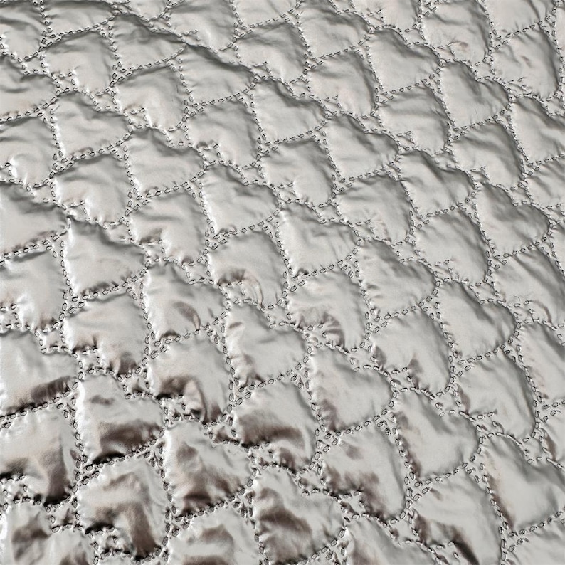 May include: Close-up of a silver quilted fabric with a repeating pattern of heart-shaped designs. The stitching is visible, creating a textured surface. The fabric has a slight sheen, reflecting light and adding depth to the design.
