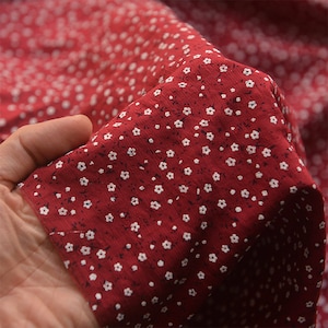 May include: Close-up of red fabric with a small white floral pattern. The fabric is held in a hand, showing the texture and drape of the material. The flowers are evenly spaced across the surface.