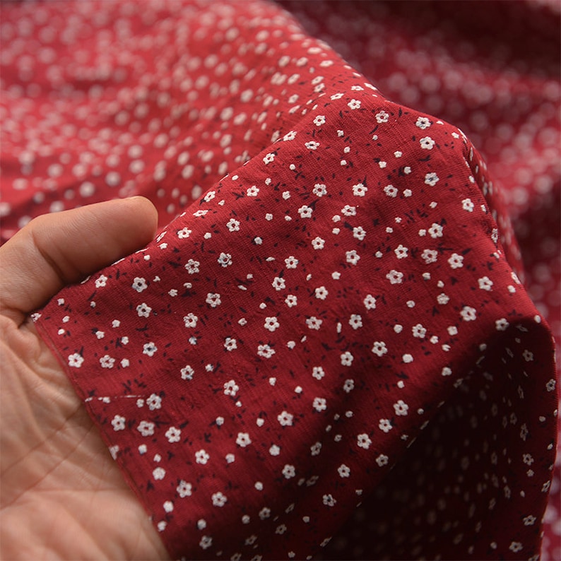 May include: Close-up of red fabric with a small white floral pattern. The fabric is held in a hand, showing the texture and drape of the material. The flowers are evenly spaced across the surface.