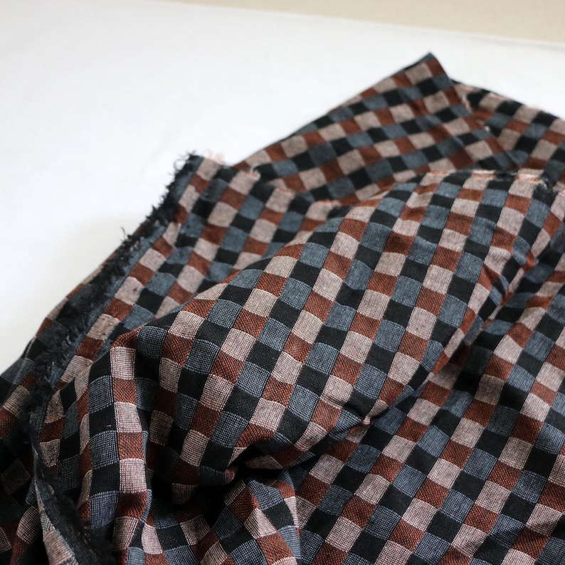 May include: Close-up of a fabric with a checkered pattern. The fabric features a repeating pattern of squares in black, brown, and gray. The fabric is folded, showing the texture and weave of the material. The edges of the fabric are frayed.
